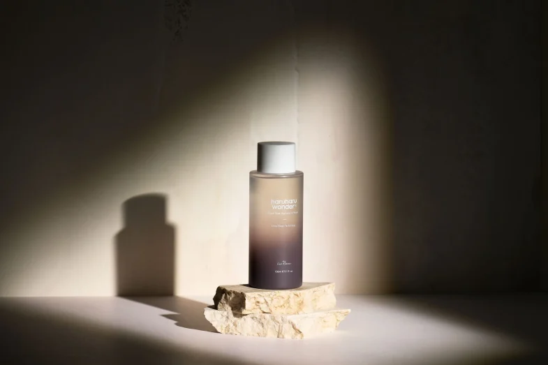 Proizvod Divote Cosmetics u stilu korejske kozmetike prikazan u minimalističkom ambijentu s fokusom svjetla na bočici koja stoji na kamenoj podlozi.
