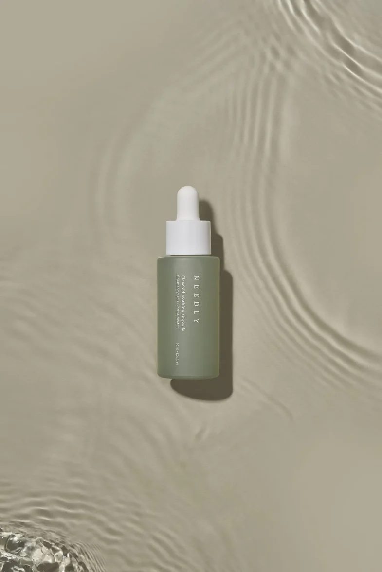 Bočica Needly seruma s pipetom nalazi se na bež površini s vodenim valovima, što predstavlja luksuzni proizvod iz korejske kozmetike, popularan u divote cosmetics.