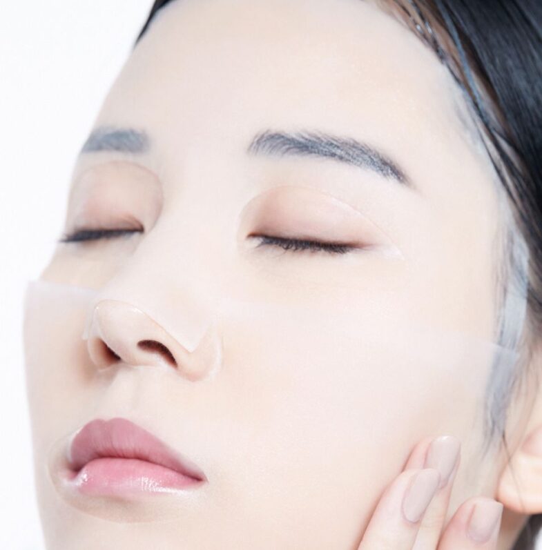 Osoba ima masku za lice koja prekriva veći dio kože, naglašavajući glatkoću i njegu lica, simbolizirajući korejsku kozmetiku i proizvode Divote Cosmetics.