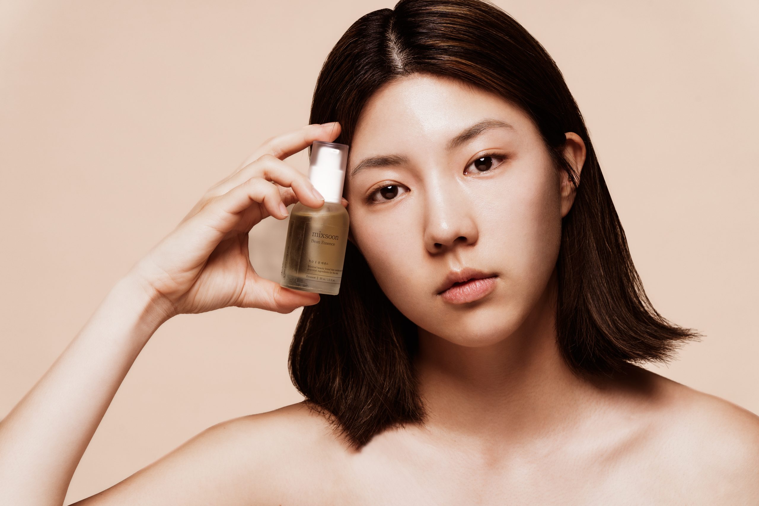 Žena s tamnom kosom drži bočicu proizvoda Divote Cosmetics iz kategorije korejske kozmetike uz lice, na bež pozadini.