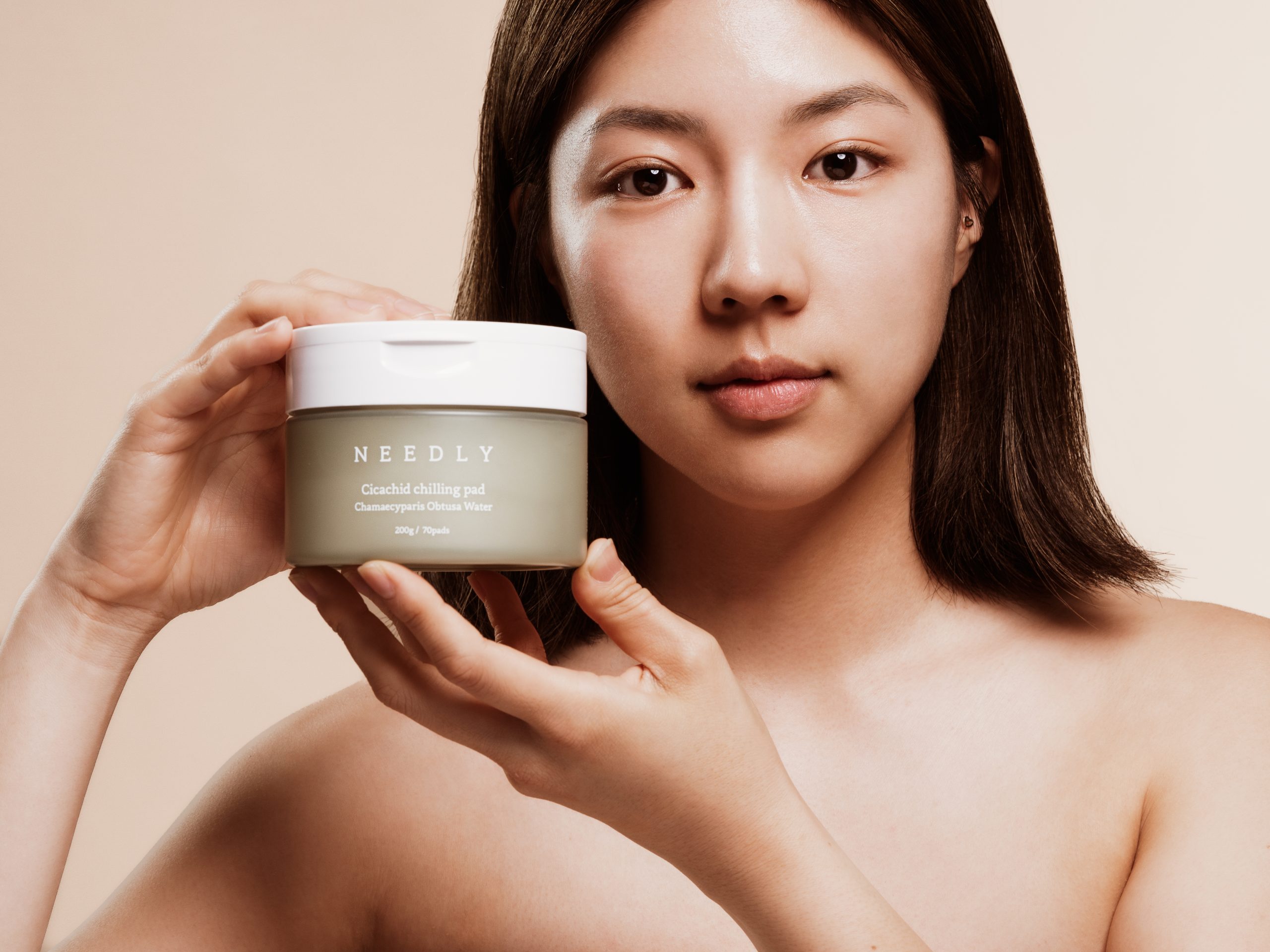 Žena drži staklenku s proizvodom iz divote cosmetics linije korejske kozmetike, označenu kao Needly Cicachid chilling pad, na neutralnoj pozadini.