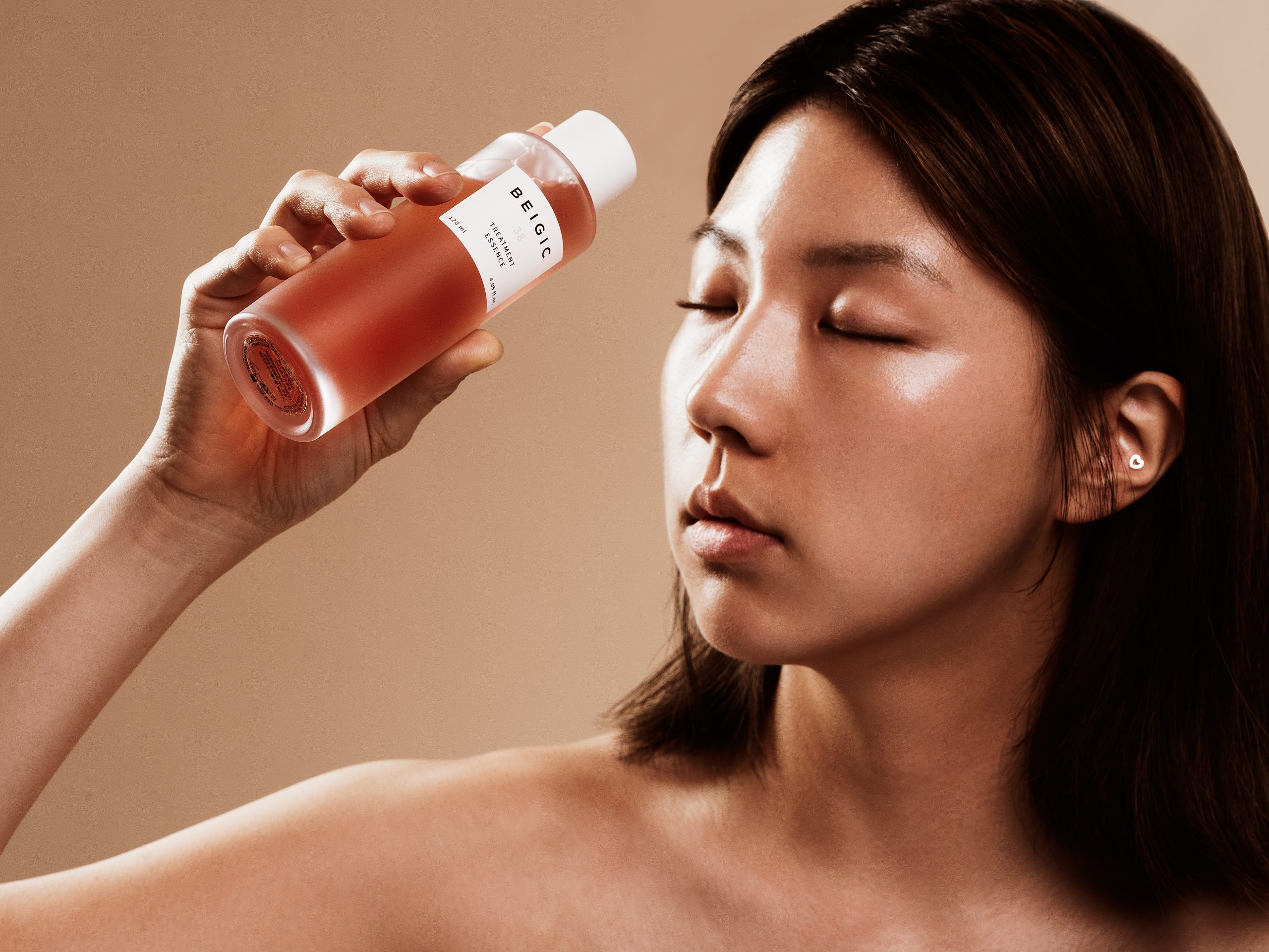 Žena zatvorenih očiju drži bocu proizvoda Divote Cosmetics, simbol korejske kozmetike, s glatkom tenom i minimalističkim dizajnom na neutralnoj pozadini.