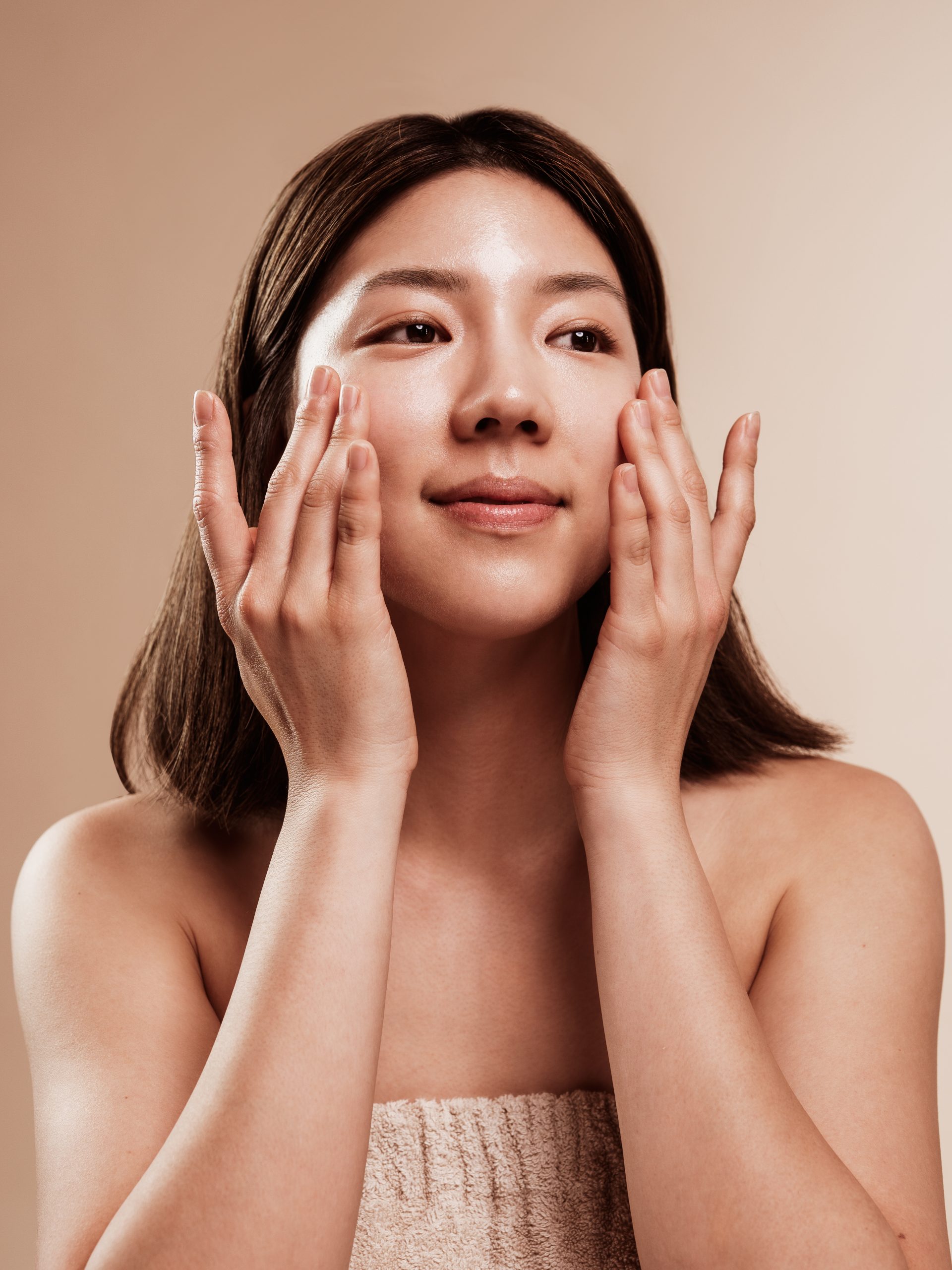 Žena se nježno dodiruje po licu, naglašavajući upotrebu korejske kozmetike i proizvoda kao što su Divote Cosmetics, na neutralnoj pozadini.