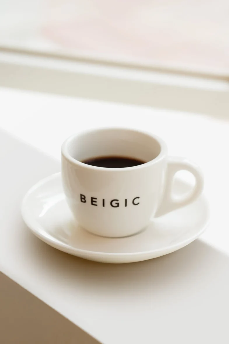 Bijela šalica s natpisom 'BEIGIC' napunjena kavom, povezana s divote cosmetics i korejskom kozmetikom, na tanjuriću uz prozor.