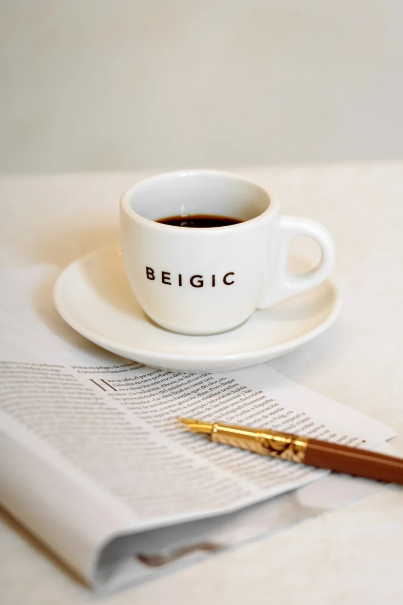 Šalica s natpisom 'Beigic' na tanjuriću, koja se povezuje s brendom Divote Cosmetics poznatim po korejskoj kozmetici, smještena na novinama uz zlatnu kemijsku olovku.