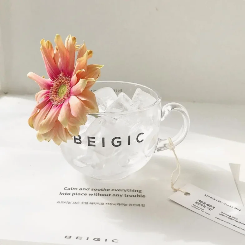 Staklena šalica s natpisom 'BEIGIC' ispunjena ledom i ukrašena cvijetom, povezana s brendom Divote Cosmetics i korejskom kozmetikom.