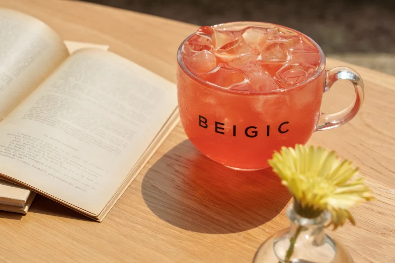 Staklena šalica s natpisom 'BEIGIC' ispunjena rozim pićem s ledom nalazi se na drvenom stolu, pored otvorene knjige i malog žutog cvijeta u vazici, podsjećajući na atmosferu povezanu s korejskom kozmetikom i divote cosmetics.