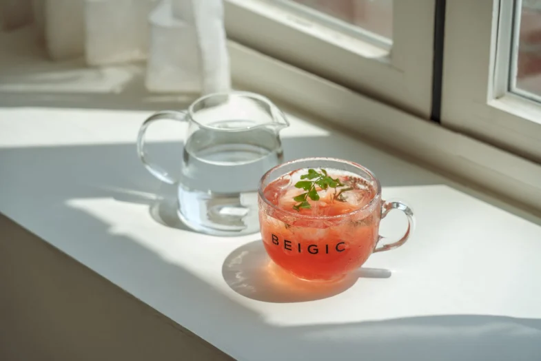 Staklena šoljica s natpisom 'BEIGIC' ispunjena crvenim napitkom s ledom i biljkama, pored staklenog vrča s vodom, smješteni na bijeloj površini ispod prozora, odražavajući atmosferu povezanosti s divote kozmetikom i korejskom kozmetikom.