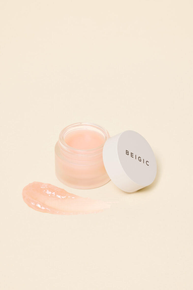 Staklena posudica s divote cosmetics proizvodom, nalik Beigic balzamu, smještena je na bež pozadini, prikazujući popularan primjer korejskih kozmetičkih proizvoda, uz prikaz guste, svijetloružičaste teksture istaknute pored posudice.