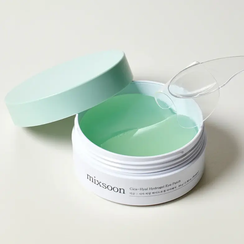 Bijela posuda s natpisom 'mixsoon' sadrži Cica-Hyal hidrogel flastere za oči, dio korejske kozmetike dostupne pod brendom Divote Cosmetics; poklopac je otvoren, a flasteri su svijetlozelene boje s prozirnim aplikatorom na vrhu.