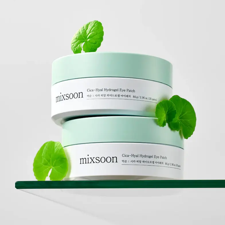 Dva pakiranja Cica-Hyal Hydrogel Eye Patch proizvoda marke Mixsoon, smještena na staklenoj površini s listovima centelle, koji ilustriraju korejsku kozmetiku i marku Divote Cosmetics.