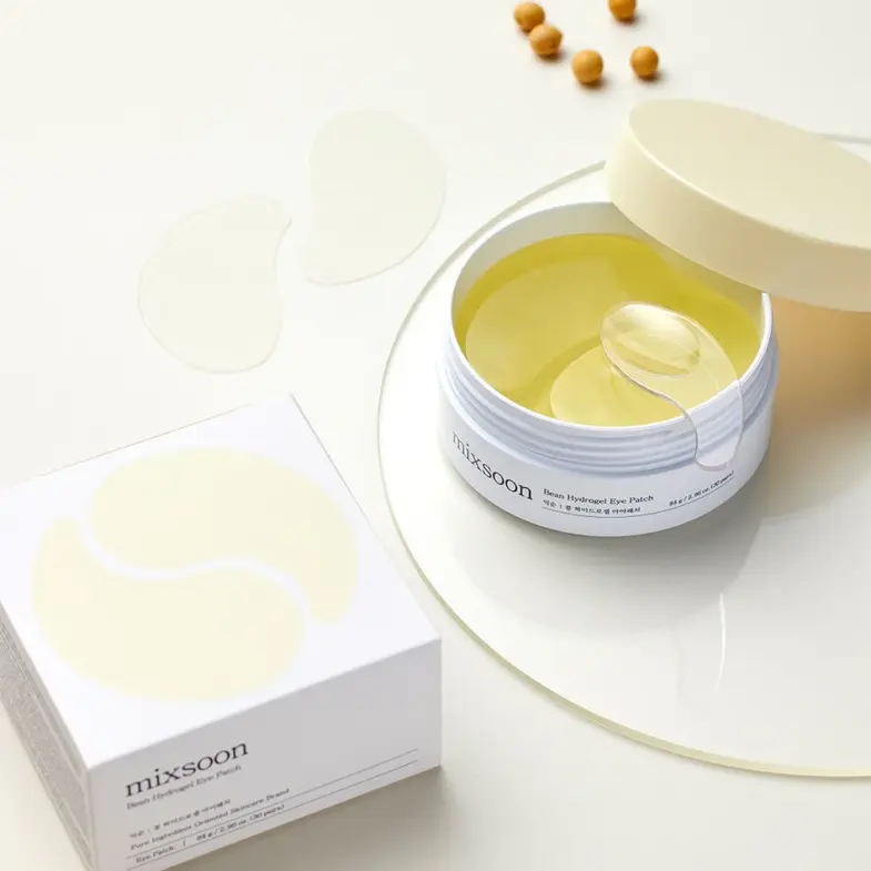 Posuda s Mixsoon hidrogel zakrpama za oči, koja pripada korejskoj kozmetici, pored kutije brenda Divote Cosmetics na svijetloj pozadini; nekoliko žutih kuglica nalazi se u blizini.