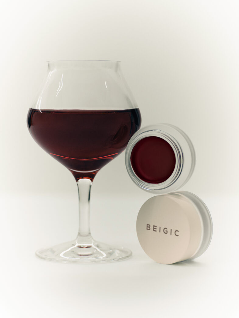 Staklena čaša crnog vina pored otvorene kutije s crvenim balzamom za usne, označene brendom BEIGIC, povezana s korejskom kozmetikom i "divote cosmetics".