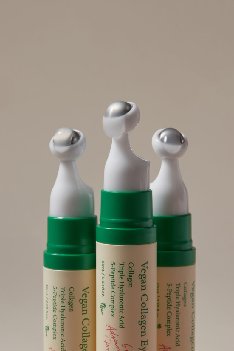 Tri proizvoda korejske kozmetike 'Divote Cosmetics' s posebnim metalnim aplikatorima na vrhu, označeni kao Vegan Collagen s hijaluronskom kiselinom i peptidnim kompleksom, stoje uspravno na neutralnoj pozadini.